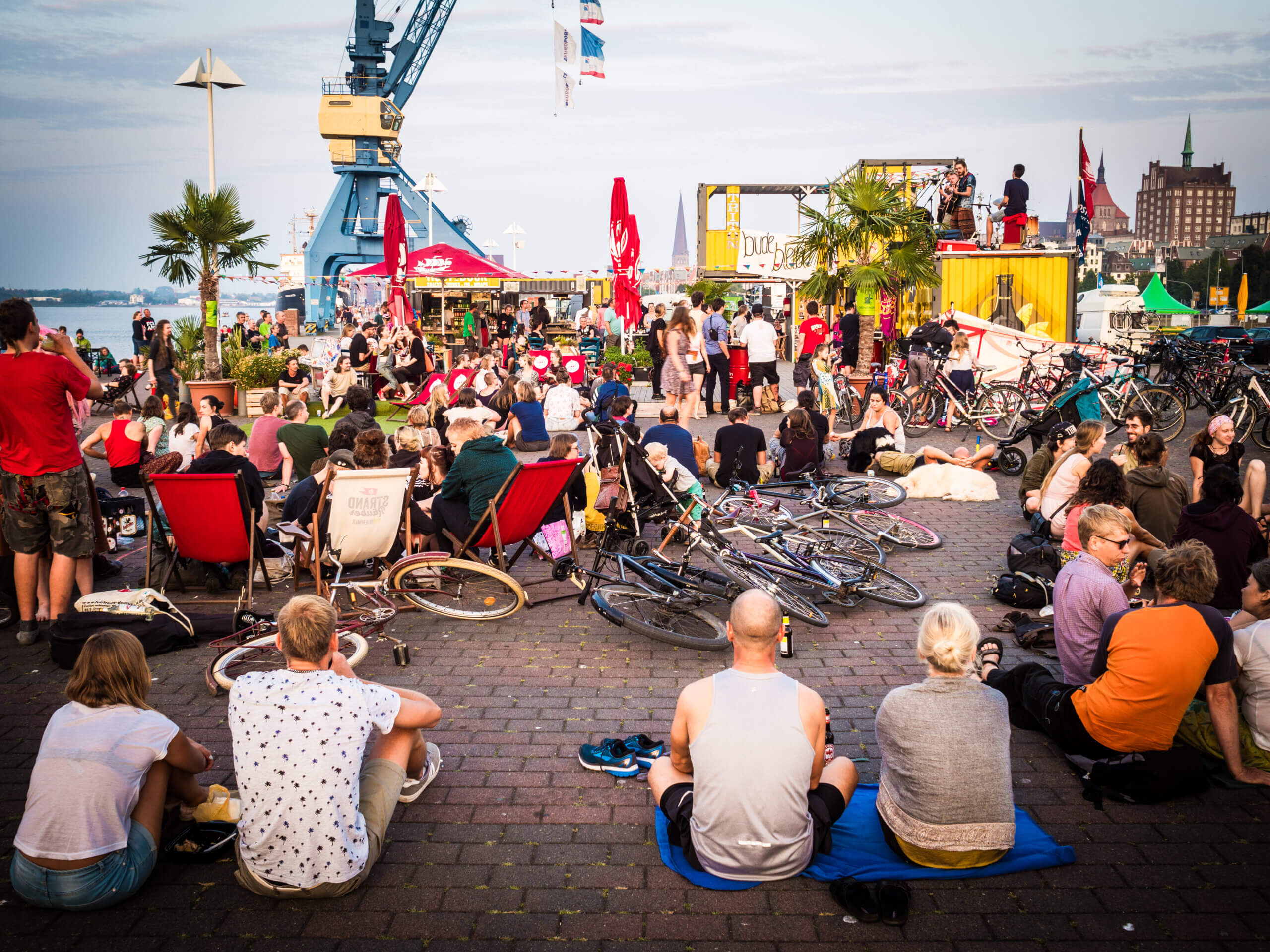 Viele Fahrräder und sitzende/stehende Menschen, im Hintergrund das Rost Dock und die Kräne