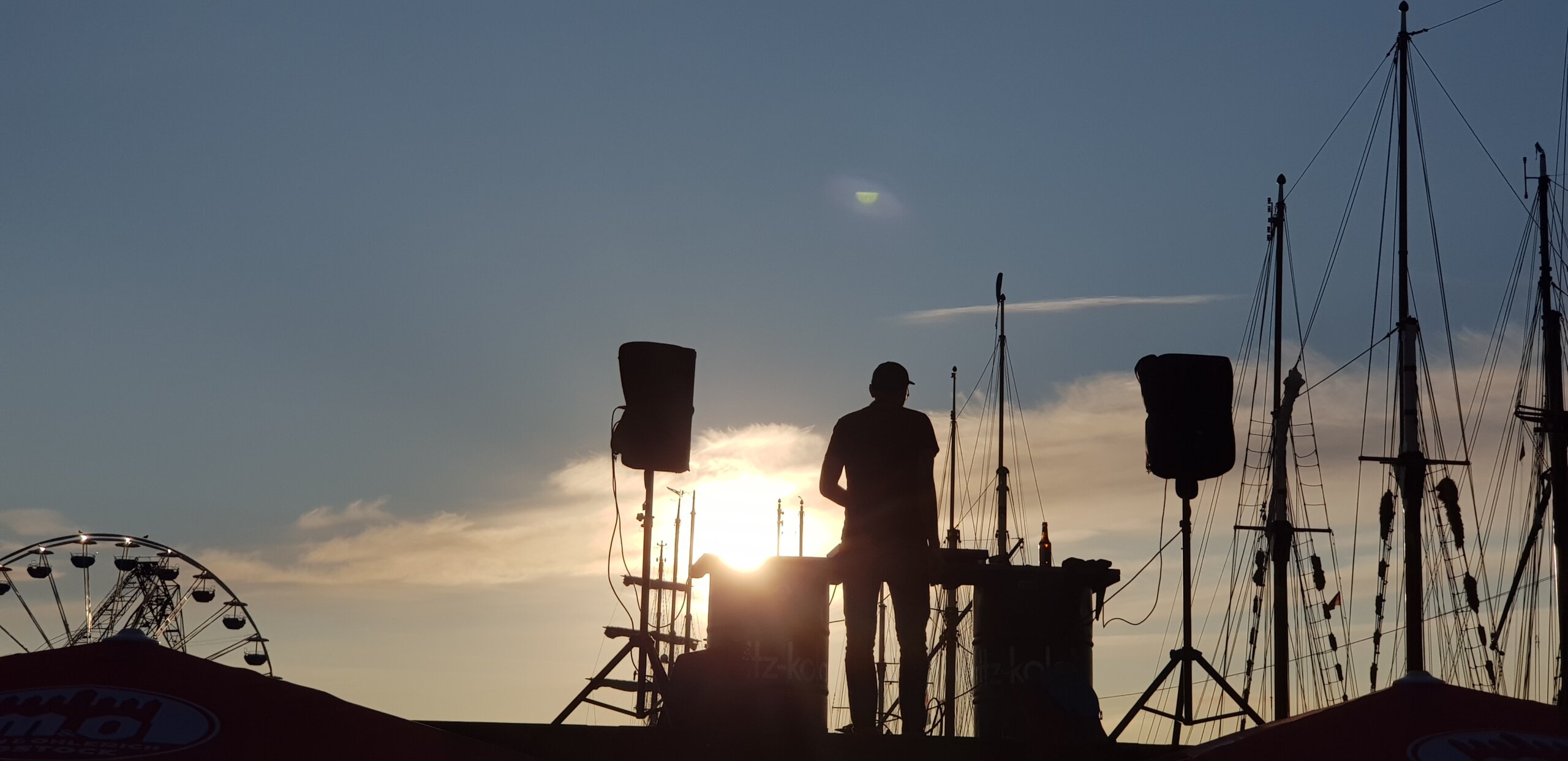 Silhouette eines DJs zwischen Boxen, im Hintergrund Segelmasten und Riesenrad